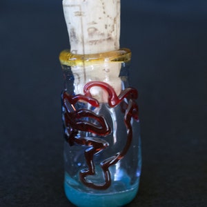 Peut inclure: Une bouteille en verre transparent avec un bouchon en li&egrave;ge. La bouteille pr&eacute;sente des motifs en verre rouge tourbillonnant et une bande jaune autour du col. La base de la bouteille est de couleur bleu clair.