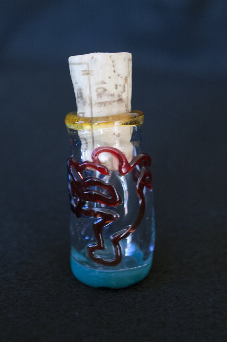 Peut inclure: Une bouteille en verre transparent avec un bouchon en li&egrave;ge. La bouteille pr&eacute;sente des motifs en verre rouge tourbillonnant et une bande jaune autour du col. La base de la bouteille est de couleur bleu clair.