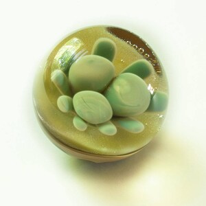 May include: A glass paperweight with a green, organic, flower-like design inside. The paperweight is clear and has a gold glitter background.