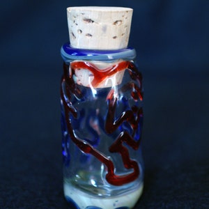 Peut inclure: Une bouteille d&eacute;corative en verre avec un bouchon en li&egrave;ge. La bouteille est transparente avec des tourbillons et des accents bleus et rouges. La base a un motif blanc et bleu. La bouteille mesure environ 10 cm de haut.