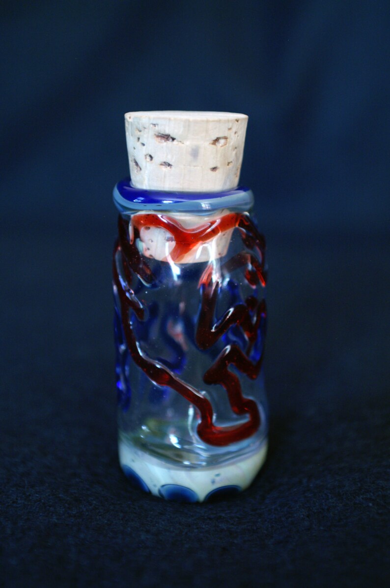 Peut inclure: Une bouteille d&eacute;corative en verre avec un bouchon en li&egrave;ge. La bouteille est transparente avec des tourbillons et des accents bleus et rouges. La base a un motif blanc et bleu. La bouteille mesure environ 10 cm de haut.