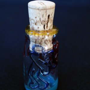 Peut inclure: Petite bouteille en verre transparent avec un bouchon en li&egrave;ge. La bouteille pr&eacute;sente des accents bleus, rouges et jaunes. Le bouchon est brun clair. La bouteille a un design unique avec des figures bleues sur le c&ocirc;t&eacute;. Le fond de la bouteille est bleu clair.