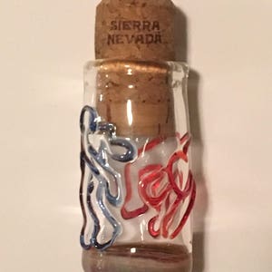 Peut inclure: Petite bouteille en verre transparent avec un bouchon en li&egrave;ge. La bouteille est orn&eacute;e de volutes rouges et bleues. Le bouchon porte l'inscription "SIERRA NEVADA". Il s'agit probablement d'un objet d&eacute;coratif ou d'un petit r&eacute;cipient.