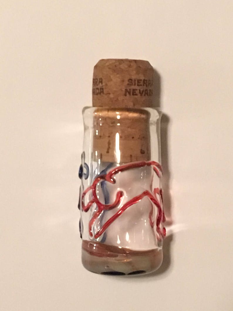 Peut inclure: Petite bouteille en verre transparent avec bouchon en li&egrave;ge. La bouteille est orn&eacute;e de volutes rouges et bleues et porte l'inscription "Sierra Nevada" sur le bouchon. La bouteille est remplie d'une substance blanche.