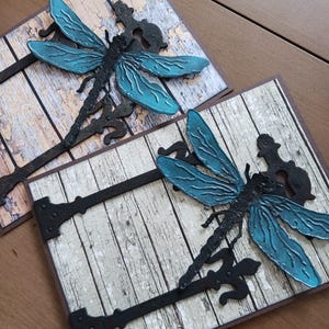 May include: Two handmade greeting cards with a rustic woodgrain background. Each card features a blue dragonfly with black accents and a metal hinge design.