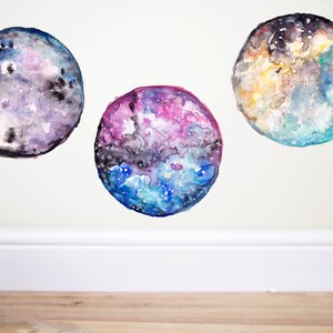 May include: Three watercolor paintings of celestial bodies. Each sphere features a unique color palette, including blues, purples, and yellows, with intricate details suggesting galaxies and nebulae. The artwork is mounted on a white wall.