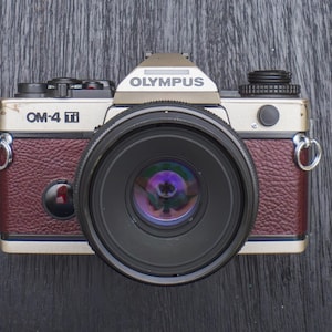 May include: Olympus OM-4 Ti camera with a large lens. The camera body is a combination of silver, burgundy, and black. The lens reflects a colorful image. The camera has the word "OLYMPUS" printed on the top.