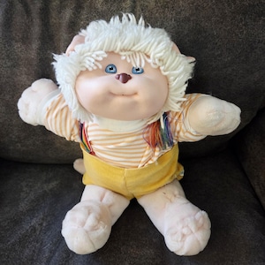 May include: A vintage plush toy with a round face, blue eyes, and white yarn hair. It wears a yellow and white striped shirt, yellow corduroy shorts, and rainbow suspenders. The toy is in a seated position with arms outstretched.