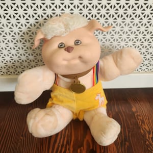 May include: A plush pink and white dog toy with a yellow and rainbow striped outfit. The toy has a gold collar with a medal and a white unicorn embroidered on the front of the outfit.