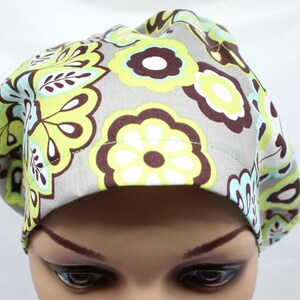 May include: A scrub cap featuring a repeating floral pattern in shades of lime green, light blue, and dark brown on a light grey background. The fabric head covering has a rounded crown and a wide, stitched band that sits across the forehead.
