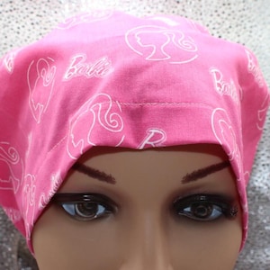 May include: A pink scrub cap featuring a repeating pattern of the word "Barbie" and the Barbie silhouette in white. The cap is made of a soft fabric and is designed to cover the head.