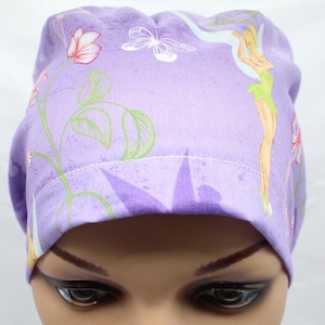 May include: A lavender scrub cap featuring a repeating pattern of a fairy with blonde hair and green clothing, white butterflies, and pink flowers with green stems. The fabric has a soft purple tone with a visible stitched hem along the front edge.