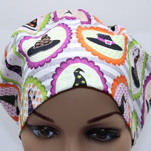 May include: A white scrub hat with a Halloween themed print featuring witch hats, pumpkins, and stars. The hat has a purple, green, and orange color scheme.