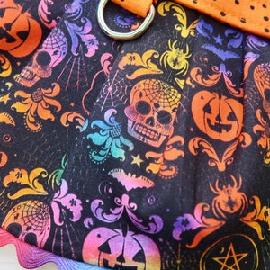 Pode incluir: Tecido preto com uma estampa arco-&iacute;ris de caveiras, ab&oacute;boras, morcegos e outras imagens com tema de Halloween. O tecido tem uma borda festonada.