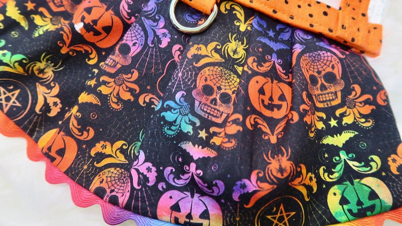 Pode incluir: Tecido preto com uma estampa arco-&iacute;ris de caveiras, ab&oacute;boras, morcegos e outras imagens com tema de Halloween. O tecido tem uma borda festonada.