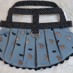 May include: A dog harness with a black and white patterned top section and a light blue skirt. The skirt features a repeating pattern of peace signs and black lips. A gold-colored metal ring is attached to the front.