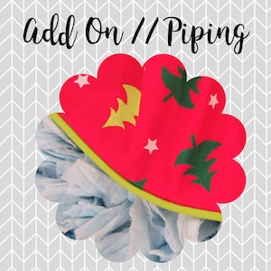 May include: A red fabric item with a scalloped edge, decorated with green leaves, yellow leaves, and white stars. The item has a lime green trim and the text "Add On // Piping" in black script.