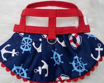 May include: Red and blue nautical themed dog harness with a white anchor and steering wheel pattern. The harness has a red adjustable strap and a silver ring.