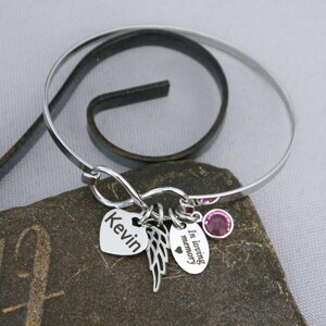 May include: A silver bangle bracelet with multiple charms. Charms include a heart with the name "Kevin", a pair of wings, and an oval charm with the words "In loving memory". A pink gemstone is set into the bracelet.