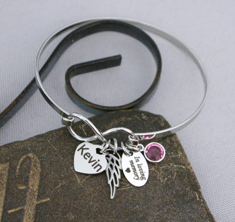 May include: A silver bangle bracelet with multiple charms. Charms include a heart with the name "Kevin", a pair of wings, and an oval charm with the words "In loving memory". A pink gemstone is set into the bracelet.