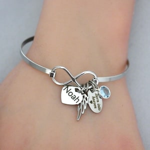 May include: A silver-toned bangle bracelet with an infinity symbol and multiple charms. Charms include a heart with the name "Noah", a feather, a small oval charm with the text "Born into heaven", and a blue gemstone.