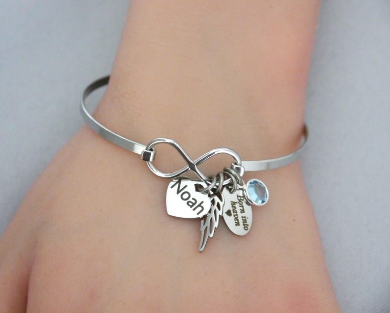 May include: A silver-toned bangle bracelet with an infinity symbol and multiple charms. Charms include a heart with the name "Noah", a feather, a small oval charm with the text "Born into heaven", and a blue gemstone.