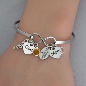 May include: Silver bangle bracelet with multiple charms. Charms include a heart with the word "Dad", a heart with the word "Mom", an infinity symbol, a small orange gem, and a charm with the words "Forever in my heart".