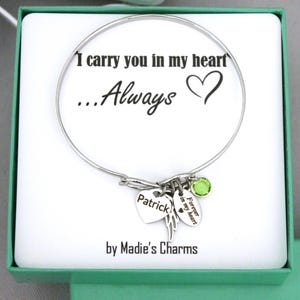 May include: A silver bangle bracelet with charms, presented in a gift box. The bracelet features a heart charm engraved with "Patrick", a wing charm, and a green gemstone. The text "I carry you in my heart...Always" is printed on the box.