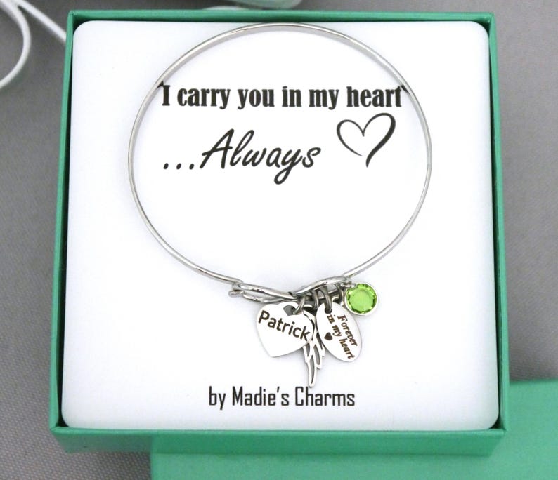 May include: A silver bangle bracelet with charms, presented in a gift box. The bracelet features a heart charm engraved with "Patrick", a wing charm, and a green gemstone. The text "I carry you in my heart...Always" is printed on the box.