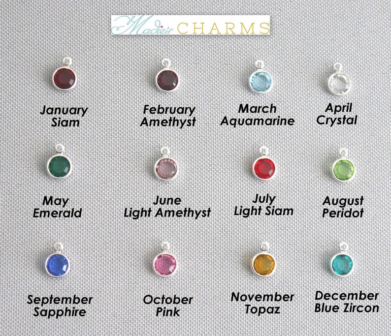 May include: A collection of silver-toned charms with various colored gemstones, including red, green, blue, and pink. Each charm is labeled with a month and gemstone name, such as "January Siam" and "September Sapphire". The brand name "Macie's CHARMS" is at the top.
