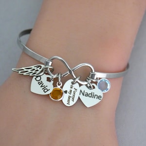 May include: Silver bangle bracelet with an infinity symbol and heart-shaped charms. Charms include a wing, a heart with "David" engraved, a heart with "Forever in my heart", and a heart with "Nadine" engraved. The bracelet also has two gemstone accents.