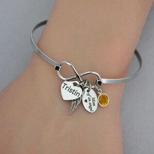 May include: A silver-toned bangle bracelet with an infinity symbol design. It features heart-shaped and oval charms, one with the name "Tristin" and another with the text "Mom of an angel." A small yellow gemstone adds a pop of color.