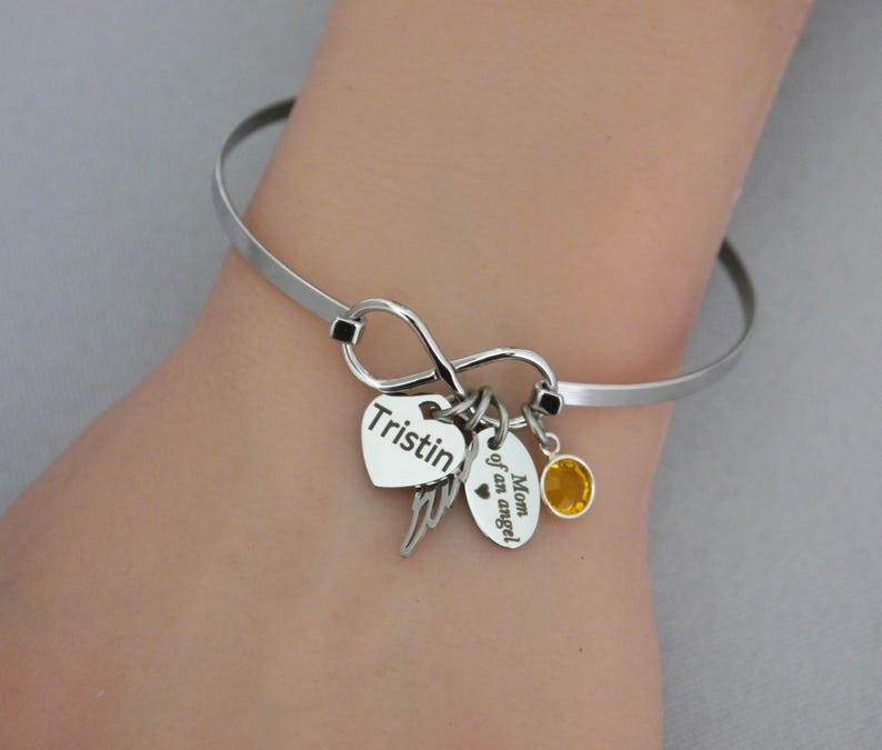 May include: A silver-toned bangle bracelet with an infinity symbol design. It features heart-shaped and oval charms, one with the name "Tristin" and another with the text "Mom of an angel." A small yellow gemstone adds a pop of color.