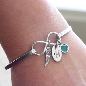 May include: Silver-toned bangle bracelet featuring an infinity symbol and three charms. Charms include a wing, a teal-colored gemstone, and an oval pendant engraved with "Forever in my heart". The bracelet is worn on a wrist.