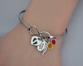 May include: A silver-toned metal bangle bracelet featuring an infinity symbol centerpiece. Attached charms include a heart with baby footprints, an oval tag engraved with the text Mommy of angels, a wing charm, and two round crystal charms in red and yellow.