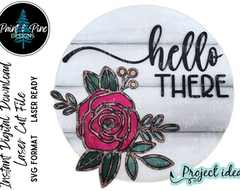 May include: A round wooden sign with a whitewashed woodgrain background. The sign features the text "hello there" in black cursive lettering. A pink and green floral design with a large rose is on the left side of the sign.