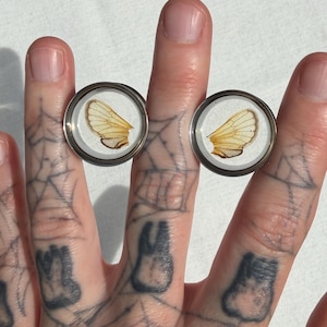 Puede incluir: Dos anillos plateados, cada uno con un ala de insecto de color beige sobre un fondo blanco. Los anillos se muestran en dedos con tatuajes intrincados, creando un contraste único entre la naturaleza y el arte corporal.
