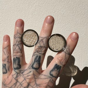 May include: Two round, silver-colored ear gauges. Each gauge features a light beige, textured center. The gauges are held up by a hand with intricate tattoos, including a spiderweb design and small, dark shapes.