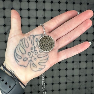 May include: A silver chain necklace with a round pendant featuring a textured, dark gray material. The pendant is held in a hand with a tattoo of a monstera leaf.