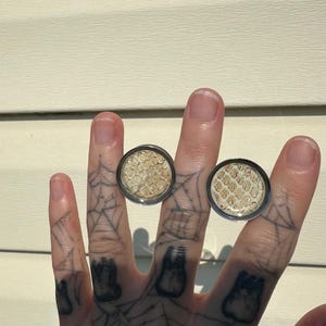 May include: Two round, silver-toned rings with a snakeskin pattern. The rings are displayed on a hand with spiderweb tattoos. The snakeskin pattern is a mix of beige and light brown.