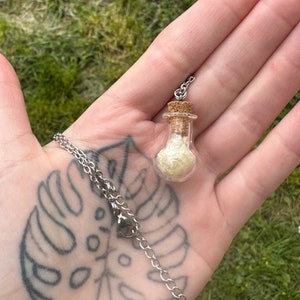 May include: A silver chain necklace with a small glass bottle pendant. The bottle is filled with a white, sparkly substance and has a cork stopper.