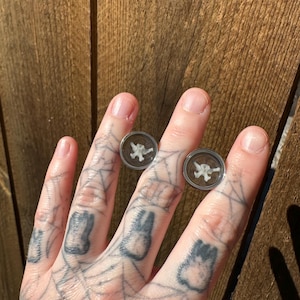 Puede incluir: Se muestran dos expansores de oreja redondos y transparentes con un diseño blanco similar a un hueso. Los expansores se colocan sobre un fondo de madera. La mano que sostiene los expansores tiene intrincados tatuajes, incluido un diseño de telaraña y varias formas.