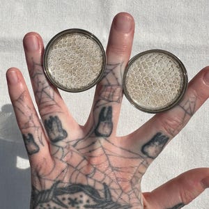 May include: Two round, silver-rimmed plugs with a textured, light-colored pattern. The plugs are held up by fingers with tattoos of teeth and a spiderweb design. The plugs appear to be for ear piercings.