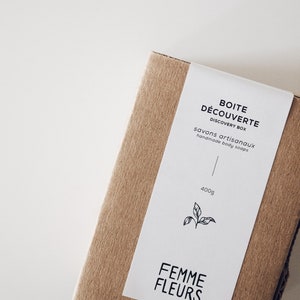 Puede incluir: Una caja rectangular de cart&oacute;n marr&oacute;n con una etiqueta blanca. La etiqueta dice "BOITE D&Eacute;COUVERTE DISCOVERY BOX savons artisanaux handmade body soaps" y "FEMME FLEURS". La caja contiene 400g de producto y una ilustraci&oacute;n de hoja.