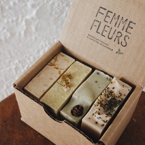 Puede incluir: Una caja de cart&oacute;n que contiene cuatro barras rectangulares de jab&oacute;n artesanal. Los jabones son de varios tonos de verde y beige, con flores secas y hierbas incrustadas. La caja est&aacute; estampada con las palabras "Femme Fleurs" en negro.