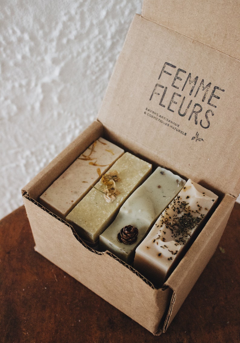 Puede incluir: Una caja de cart&oacute;n que contiene cuatro barras rectangulares de jab&oacute;n artesanal. Los jabones son de varios tonos de verde y beige, con flores secas y hierbas incrustadas. La caja est&aacute; estampada con las palabras "Femme Fleurs" en negro.