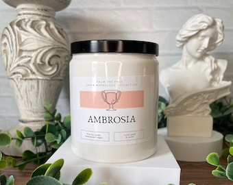 May include: White candle jar with a black lid. The label says "From the Page Greek Mythology Collection" and features a gold cup with a pink background. The candle is labeled "Ambrosia" and has the scents "Honey, Peach & Vanilla".