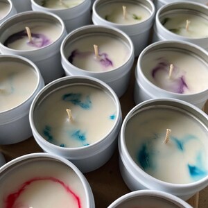 May include: A collection of small, white, round candle tins with colorful wax inside. The candles have a single wick in the center and are arranged in a random pattern.