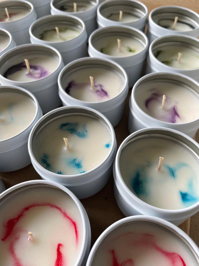 May include: A collection of small, white, round candle tins with colorful wax inside. The candles have a single wick in the center and are arranged in a random pattern.