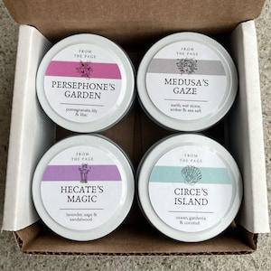 May include: Four small round metal tins with lids, each featuring a different design and scent. The tins are arranged in a 2x2 grid. The top left tin is labeled "Persephone's Garden" with the scent "pomegranate, lily & lilac". The top right tin is labeled "Medusa's Gaze" with the scent "earth, wet stone, amber & sea salt". The bottom left tin is labeled "Hecate's Magic" with the scent "lavender, sage & sandalwood". The bottom right tin is labeled "Circe's Island" with the scent "ocean, gardenia & coconut".